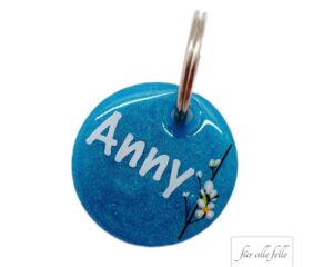Runde personalisierte Hundemarke mit dem Namen 'Anny' in Weiß auf einem glitzernden blauen Hintergrund mit einem zarten Blumenmuster, handgefertigt von 'Für alle Felle'.